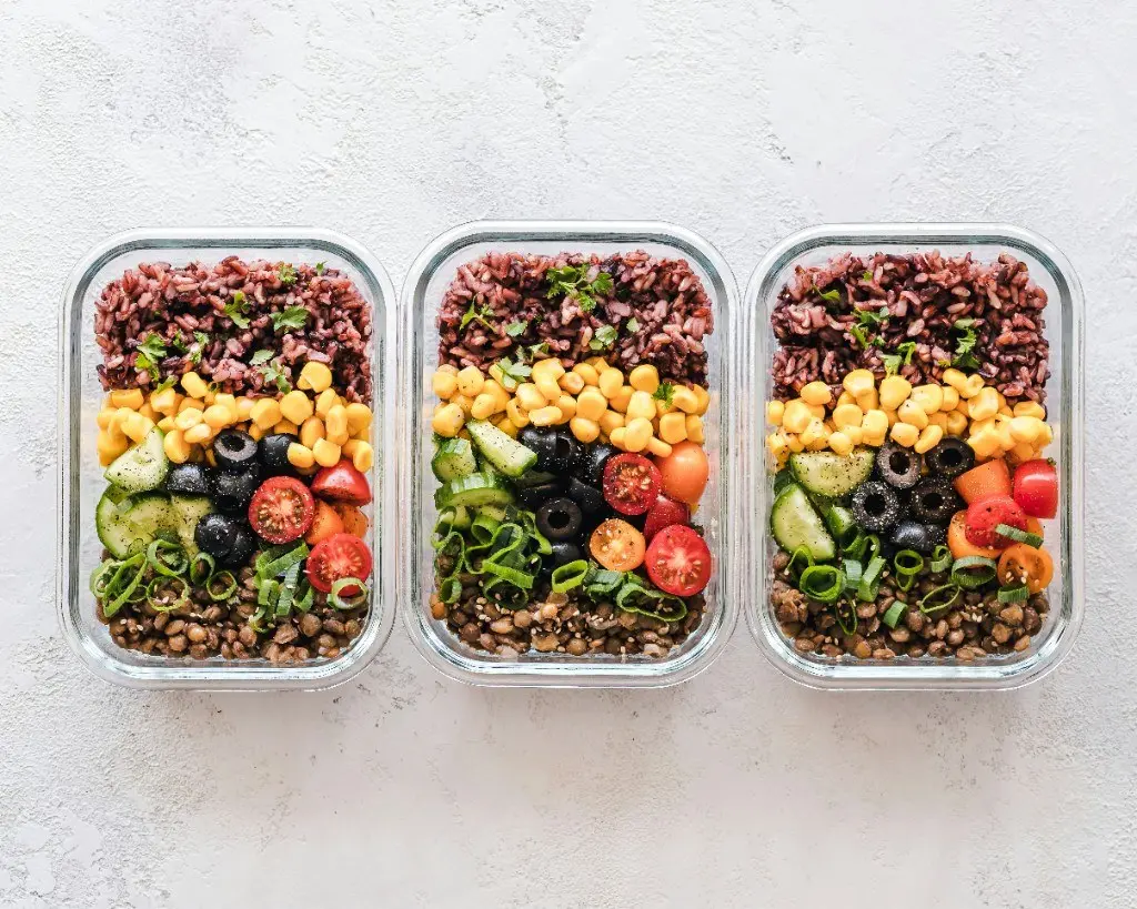 Glass meal prep containers filled with rice, lentils, corn, olives, tomato, and cucumber